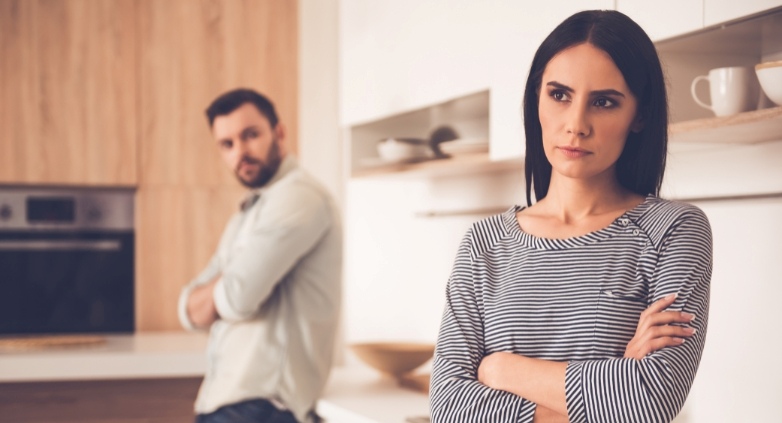 A man and woman in a kitchen, arms crossed, represent the emotional and relationship signs indicating a potential drinking problem.
