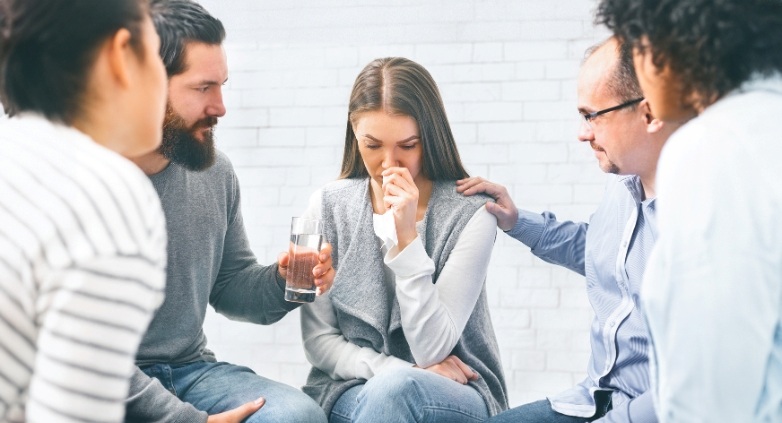 Several individuals gathered around a table, one person holding a glass, focused on discussing addiction treatment options.