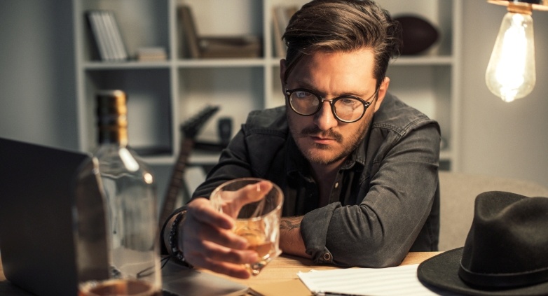 A bespectacled man at a desk uses a laptop, accompanied by a glass of whiskey, exploring the topic of familial alcoholism.