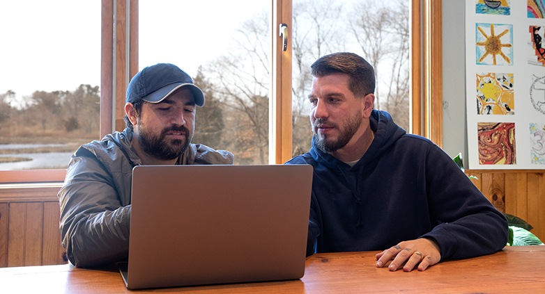 Two men sit at a wooden table, focused on a laptop. One wears a cap and jacket, the other a hoodie.
