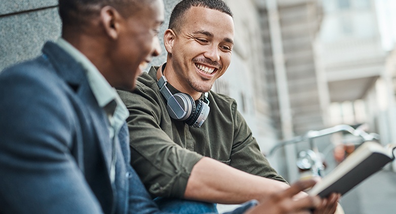 Two men sit outdoors, smiling and engaged in conversation. One wears headphones around his neck and holds a book, creating a friendly, relaxed atmosphere.