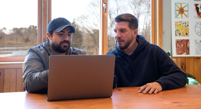 Two men at a table with a laptop, engaged in a conversation about encouraging a family member to seek addiction treatment.