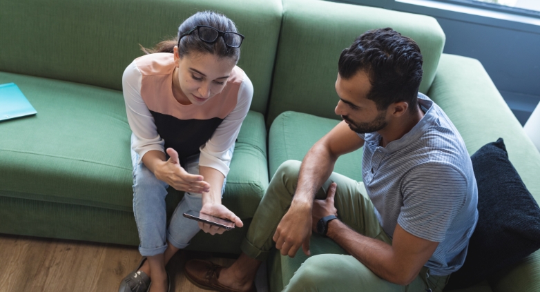 A man and woman sit on a couch, focused on a mobile phone, engaging in an online support group for families of addicts.