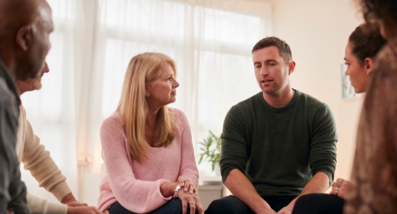 A group of six people is engaged in a discussion in a bright, cozy room. They appear attentive and supportive.