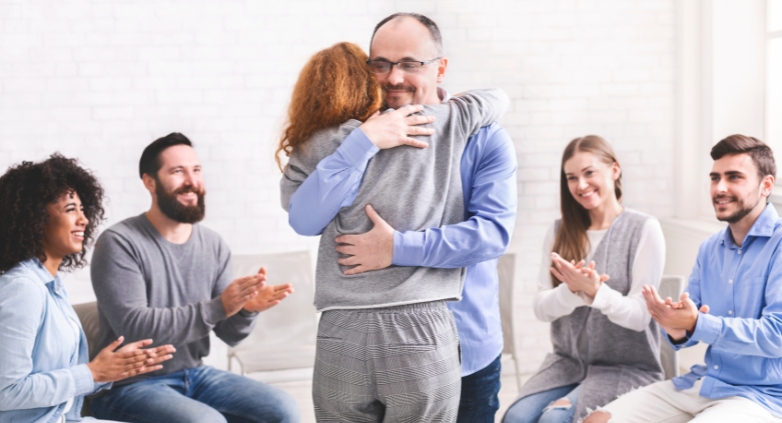 A man embraces a woman, surrounded by a supportive group, highlighting the benefits of family therapy for addiction treatment.