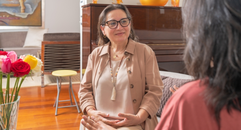 In a living room, one woman talks to another about ways to assist an alcoholic mother, focusing on helpful steps.