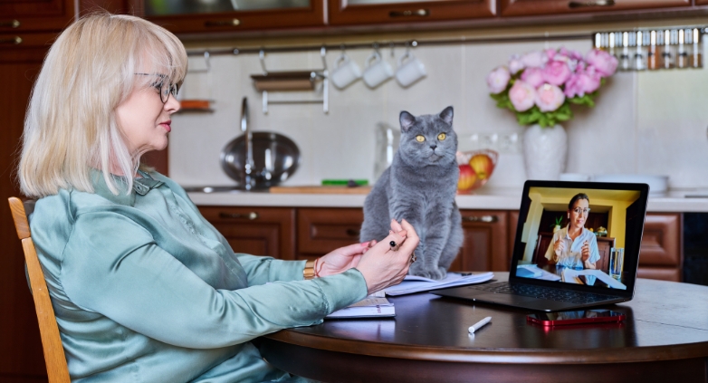 A woman sitting at a table with a cat on the table, focused on a virtual meeting about supporting an alcoholic mother.