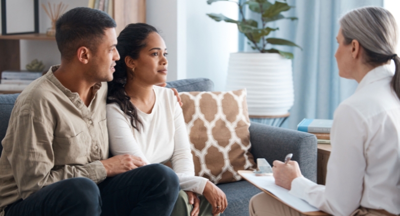A couple sits on a couch, talking to a therapist about the difficulties families face in addiction recovery and support.