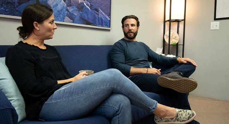 A man and woman are seated on a couch in an office, engaged in a conversation about support groups for families of addicts.