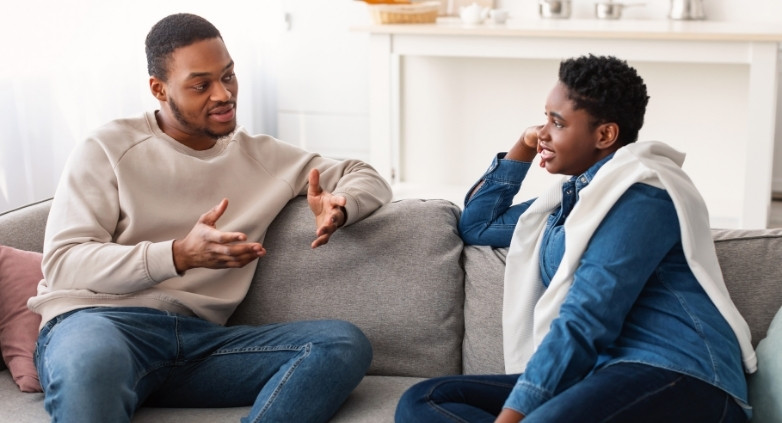 A couple sits on a couch, engaged in a serious conversation about mental health struggles related to childhood experiences.